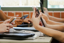 Học sinh trung học ngồi ở bàn cầm nhiều thiết bị kỹ thuật số như điện thoại và máy điện toán bảng trong giờ học. (Ảnh: LBeddoe/Shutterstock)